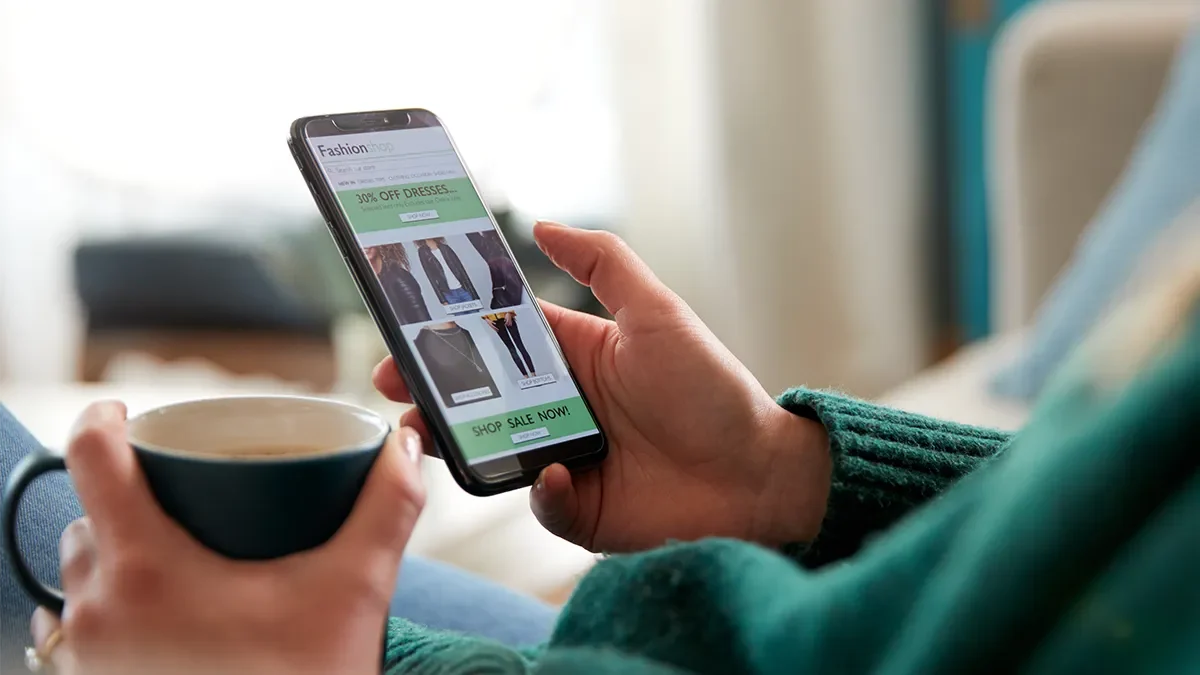A person wearing green holds a phone and a coffee as they browse online shops, without completing a purchase. Expert digital marketing advice from Webmart.