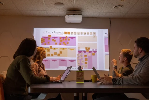 A team of Webmarteers sit around a table with a screen showing a moodboard of post it notes as they brainstorm marketing strategies for clients.