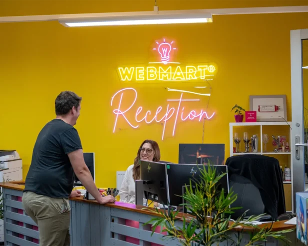 Two Webmarteers smile at each other as one stands and one sits behind the reception desk in our Yellow Shed HO in Bicester.