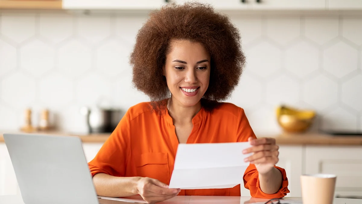 A woman in an orange top smiles as she reads a piece of direct mail at her breakfast table, with her laptop to one side and coffee to the other